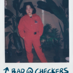 orono wearing a red jumpsuit surrounded by fake plants, taken from the set of the on & on music video shoot. the caption says 'bad at checkers.'